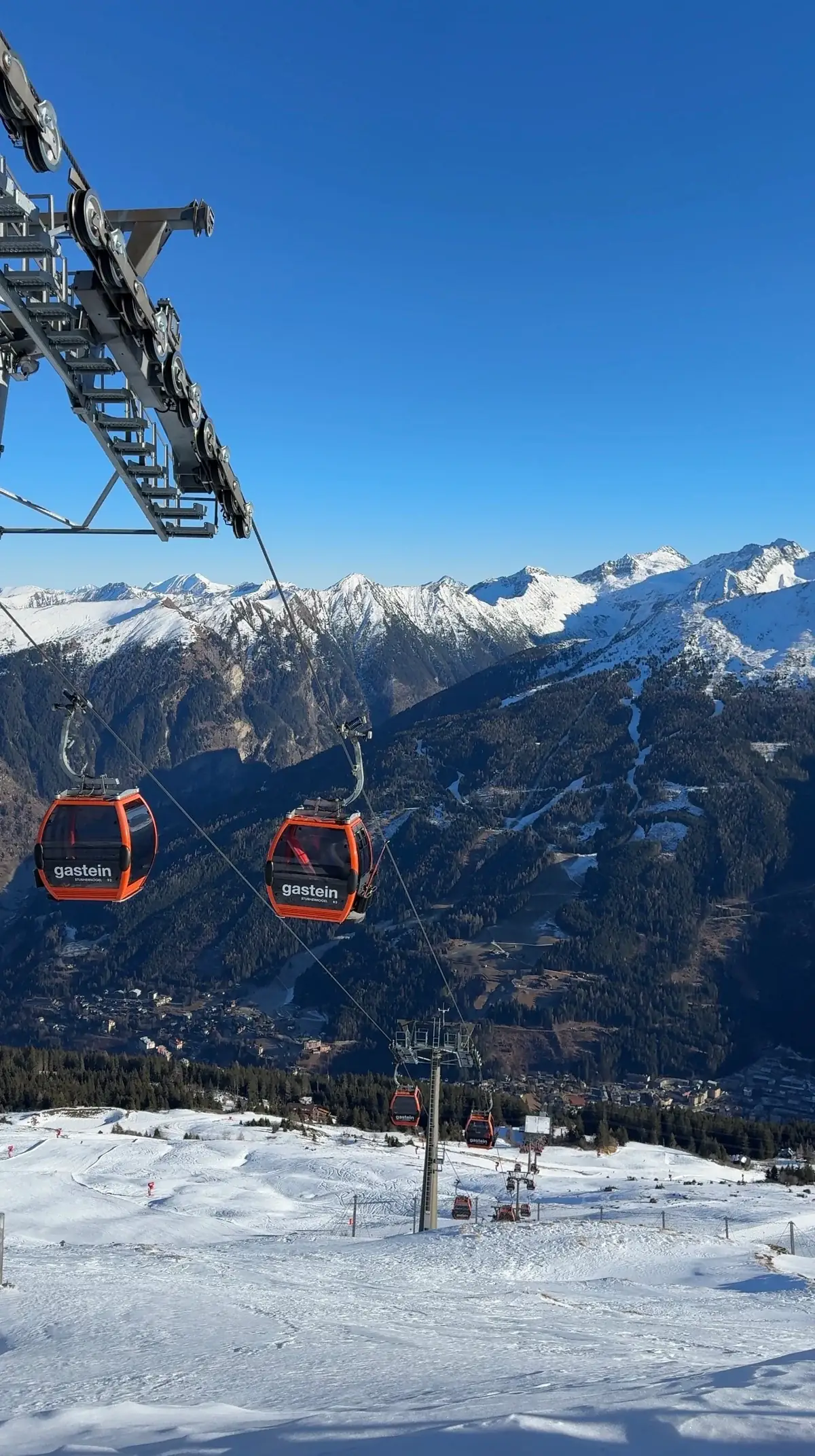 Red Gastein gondolas above a snowy slope, with a valley and mountain ridges in the background.