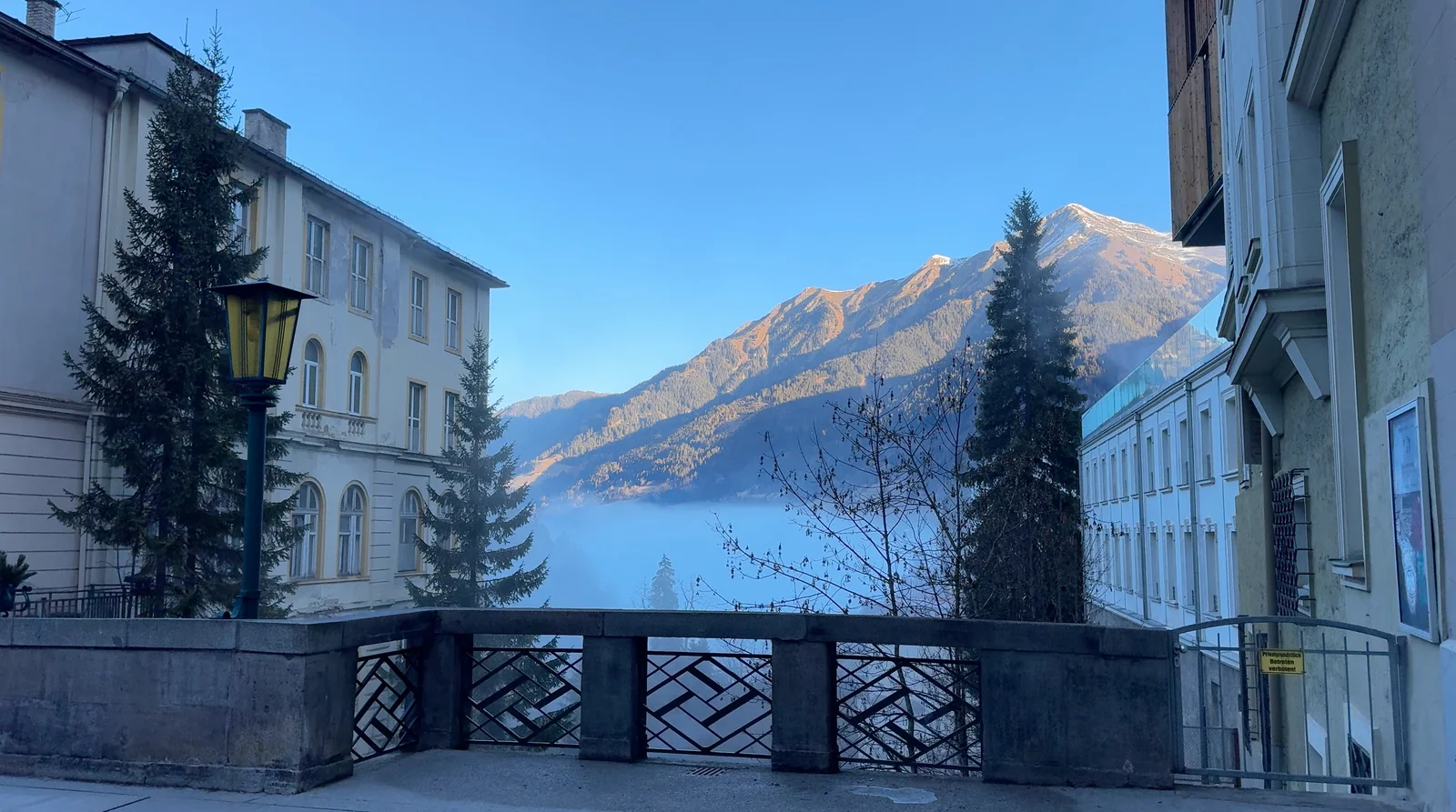 A bridge railing beside the waterfall, with town buildings, mist and a mountain ridge beyond.