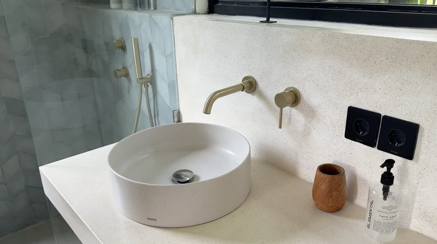 A bathroom with a round white basin, brass taps and a shower behind