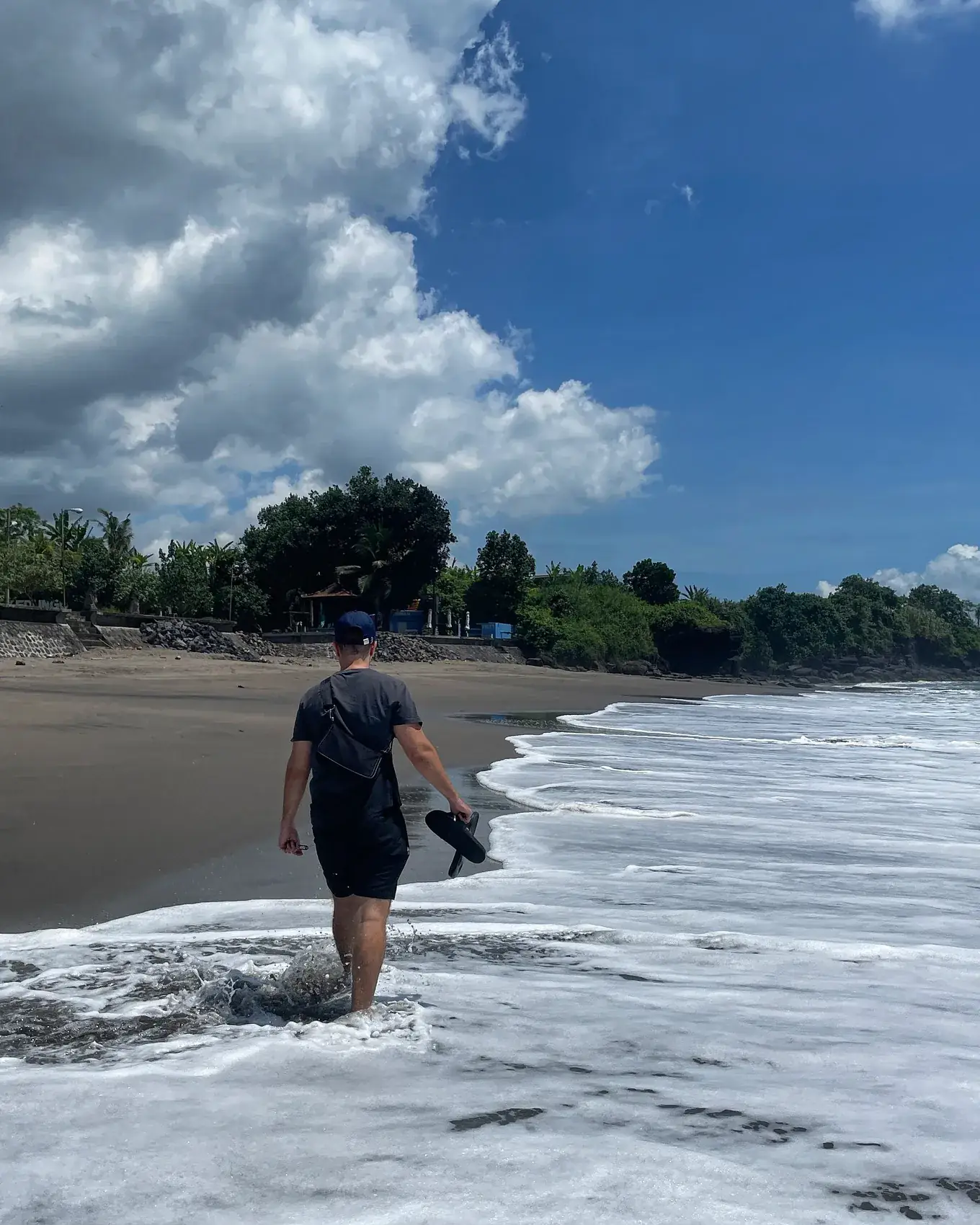 A man walking through the foam on a dark sand beach under a blue sky