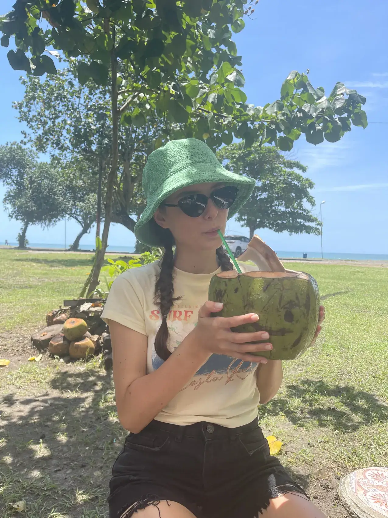Me in a green bucket hat holding a fresh coconut under a tree near the sea
