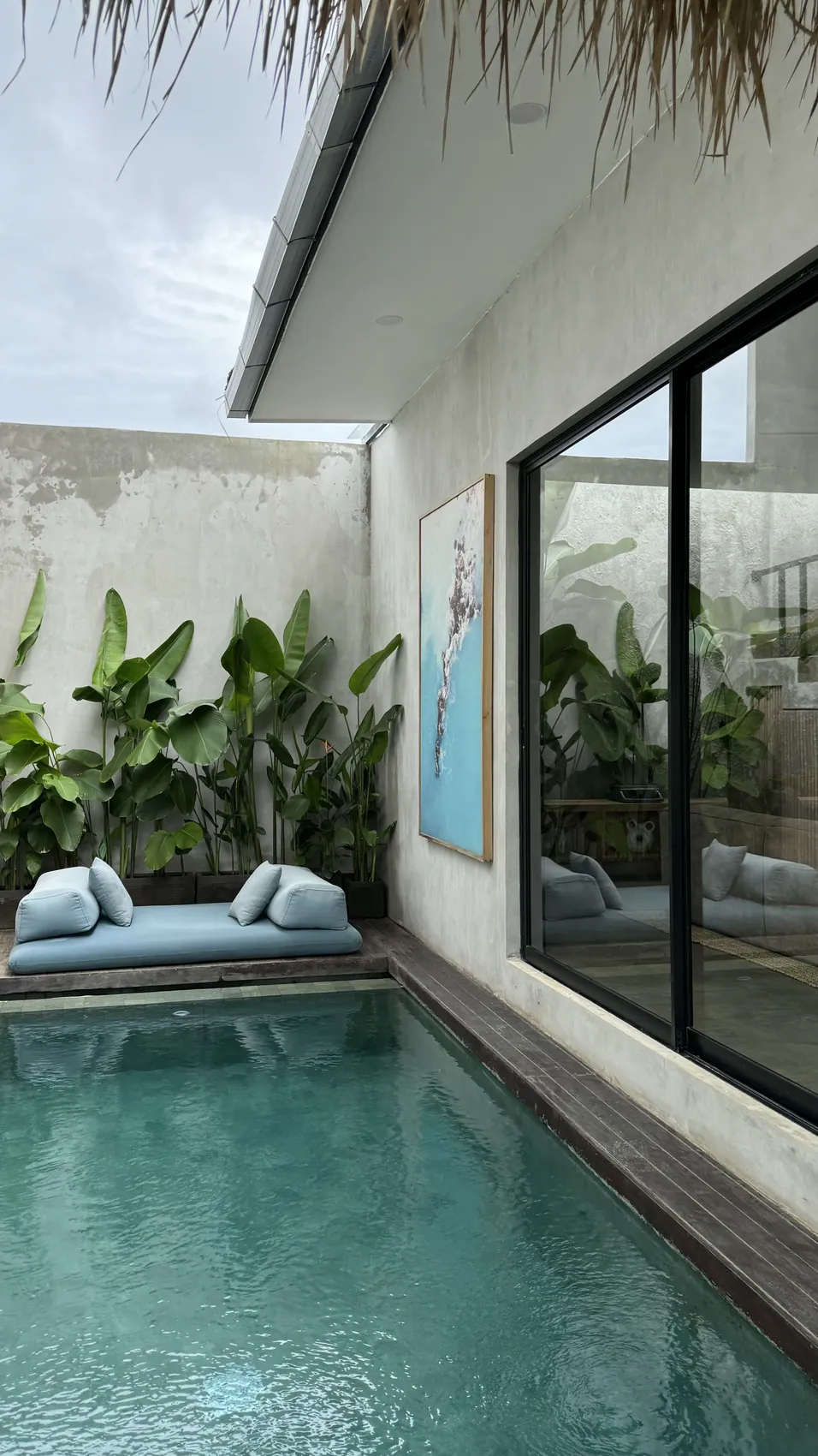 A pool with a daybed and tropical plants next to a villa with a thatch roof