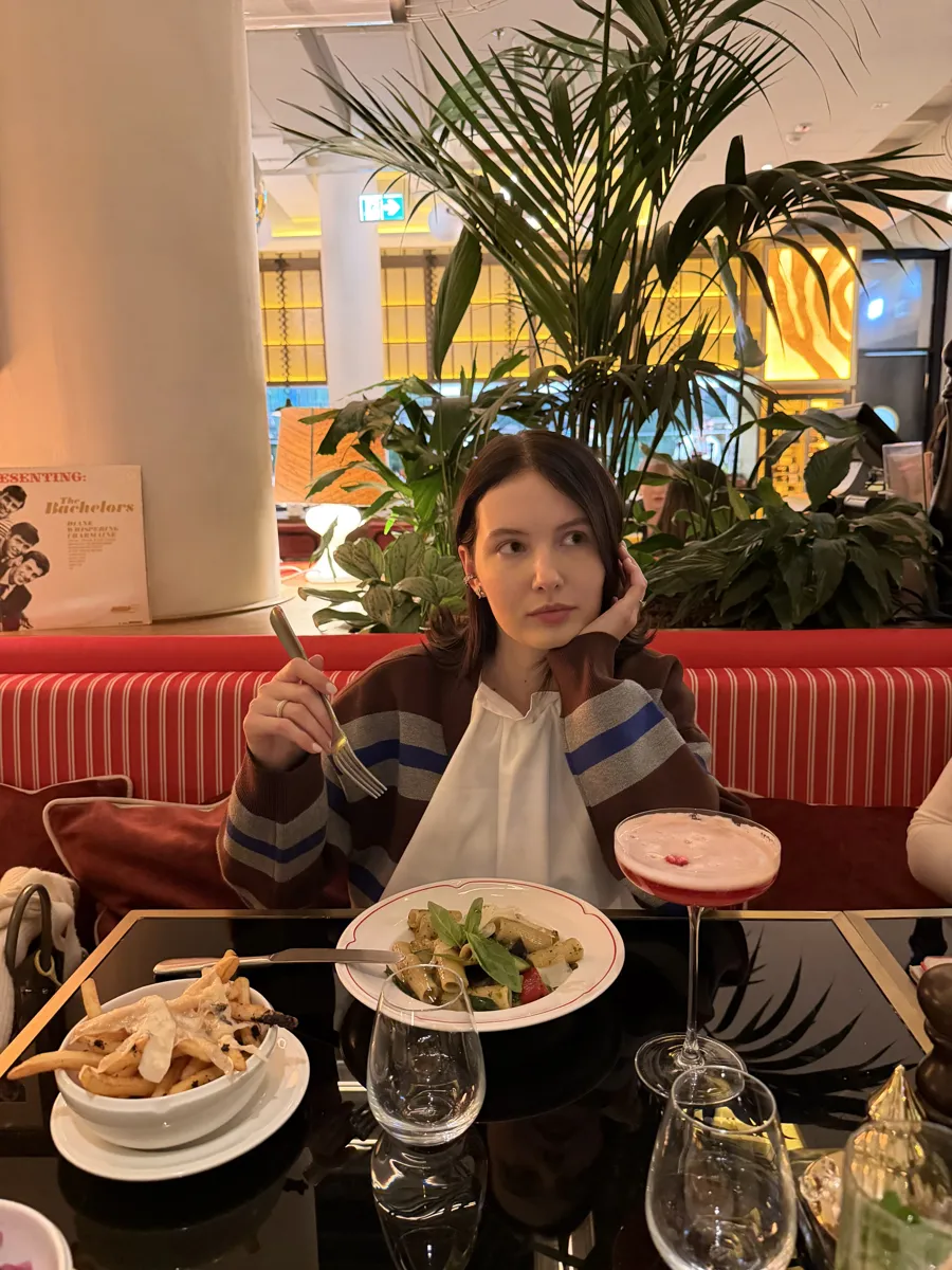 A woman seated at a red banquette with plates of pasta and fries on the table.