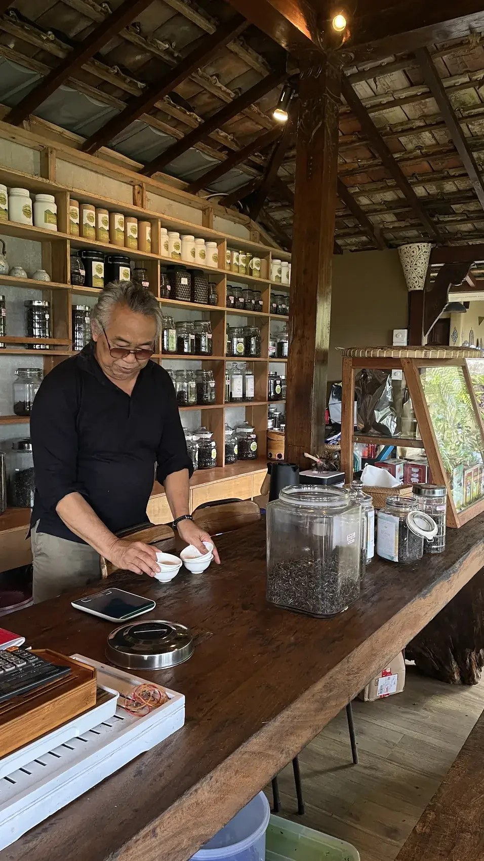 A tea master in black preparing tea at a long wooden table in a hall with shelves of tea