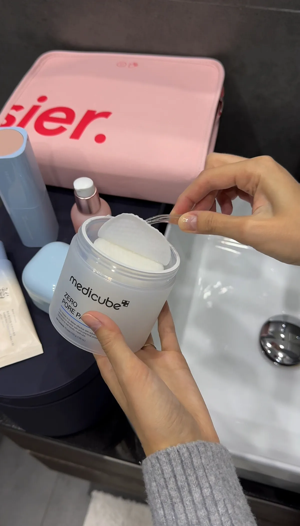 A hand lifts a white round pad with plastic tongs out of an open Medicube Zero Pore Pad 2.0 jar beside a bathroom basin.