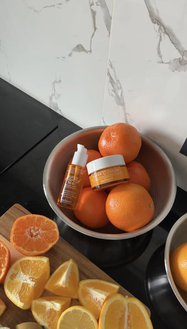 An orange Melumé jar and bottle in a bowl of oranges on a marble counter.
