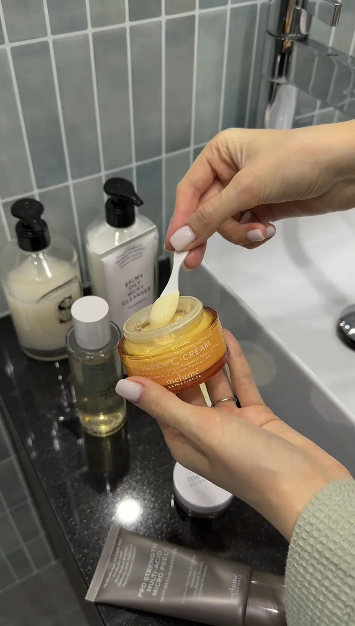 A hand scooping cream from an orange Melumé jar with a small spatula.