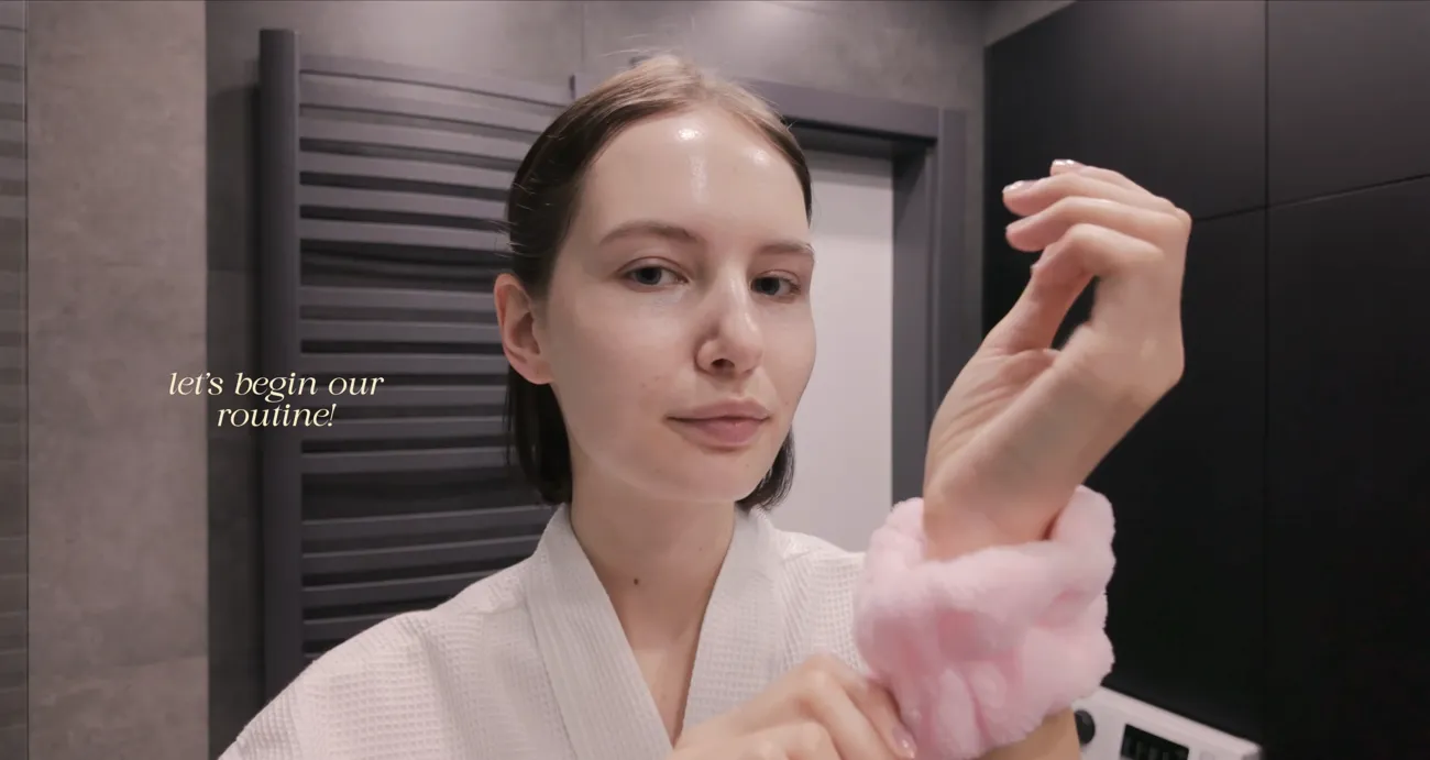 A woman in a pale pink robe and a pink wrist scrunchie at the start of a skincare video.