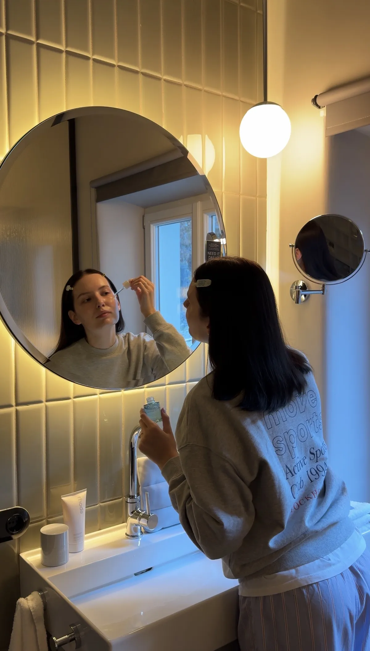 A woman in a grey sweatshirt applies a skincare product with a dropper in front of a round bathroom mirror.