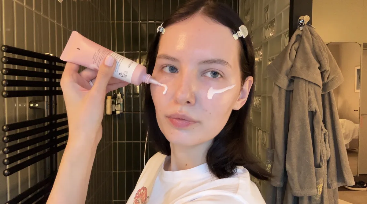 A woman applies a line of sunscreen from a pink tube to her cheek in a green-tiled bathroom.