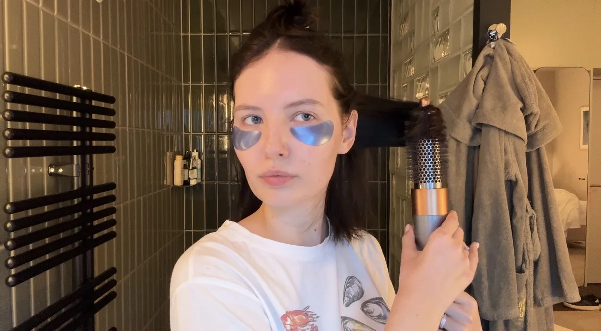 A woman with dark blue eye patches under her eyes styles her hair with a Dyson Airwrap in a green-tiled bathroom.