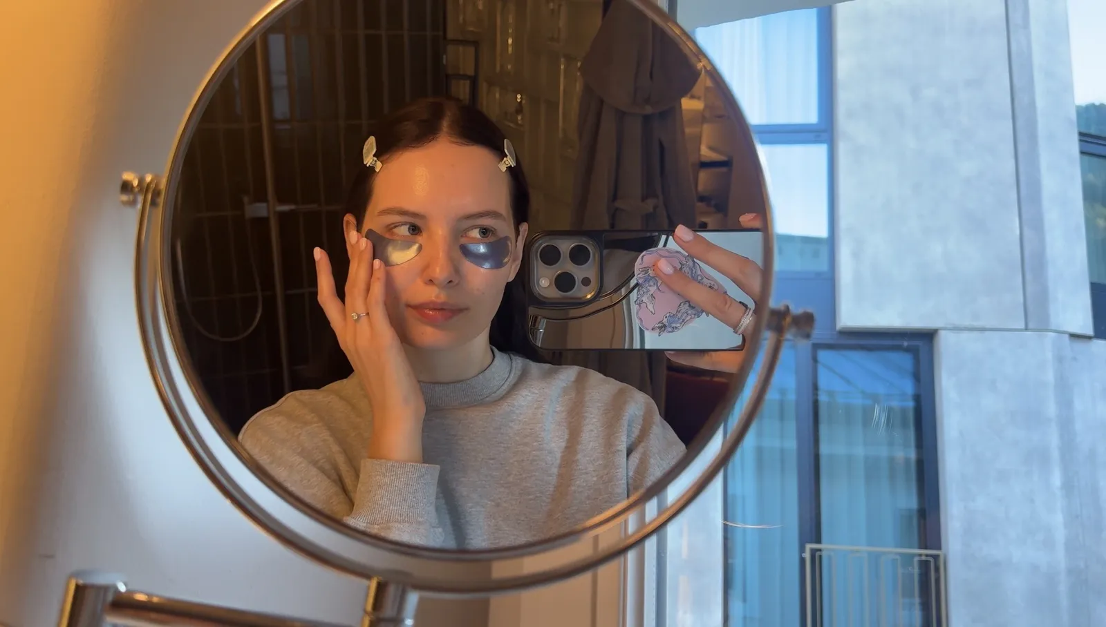 A woman in a grey sweatshirt touches the skin near her eye patches while looking into a round mirror.