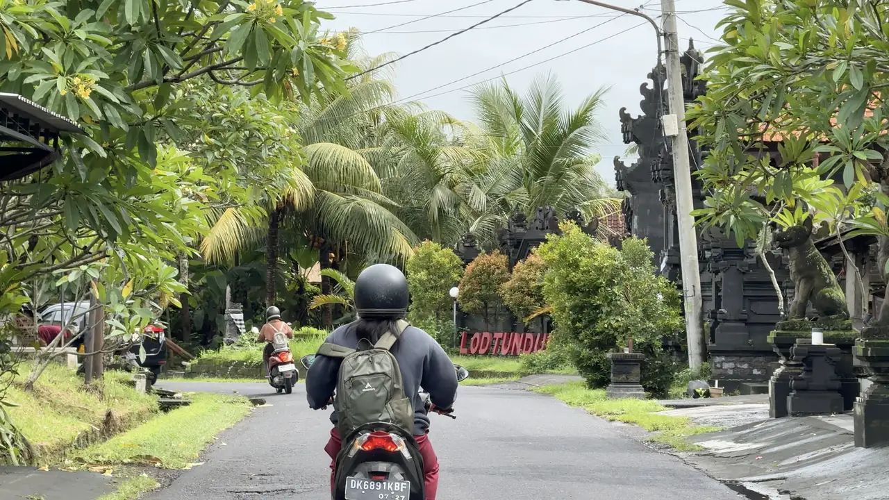 A scooter rider with a backpack on a road lined with palm trees and a Balinese gate