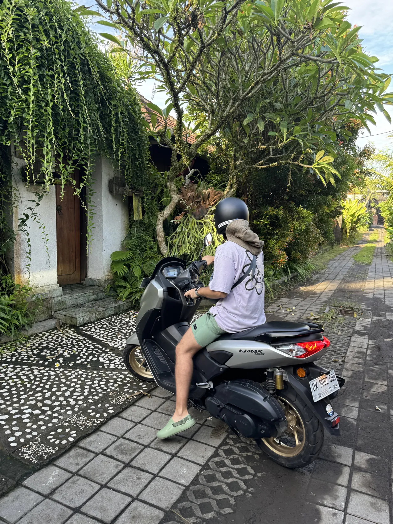 A person in a helmet sitting on a parked Yamaha NMAX scooter in a quiet lane lined with plants