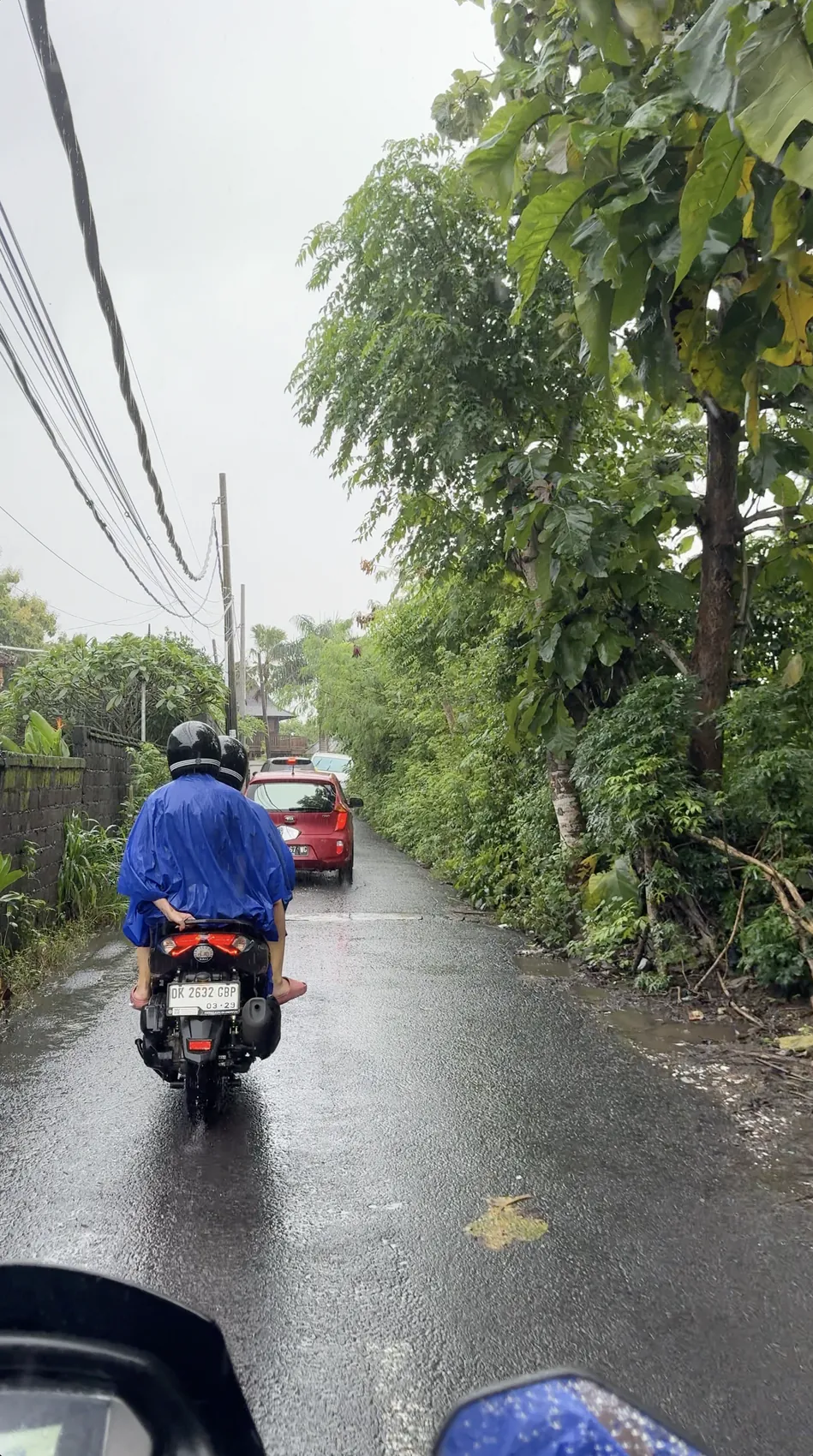 A person in a blue raincoat riding a scooter along a wet narrow road
