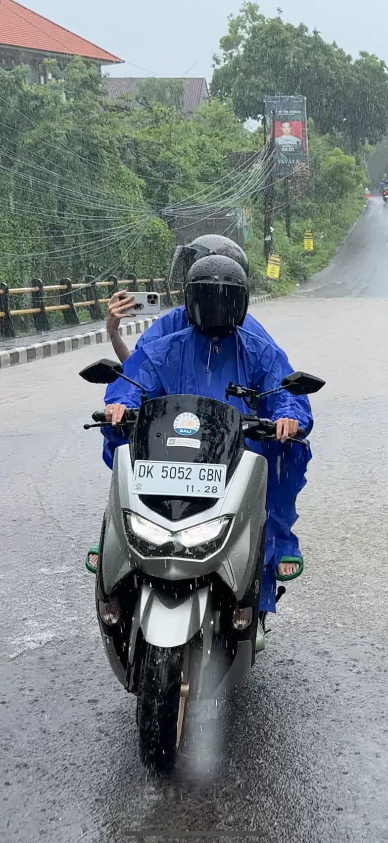 A person in a blue raincoat riding a scooter along a wet road