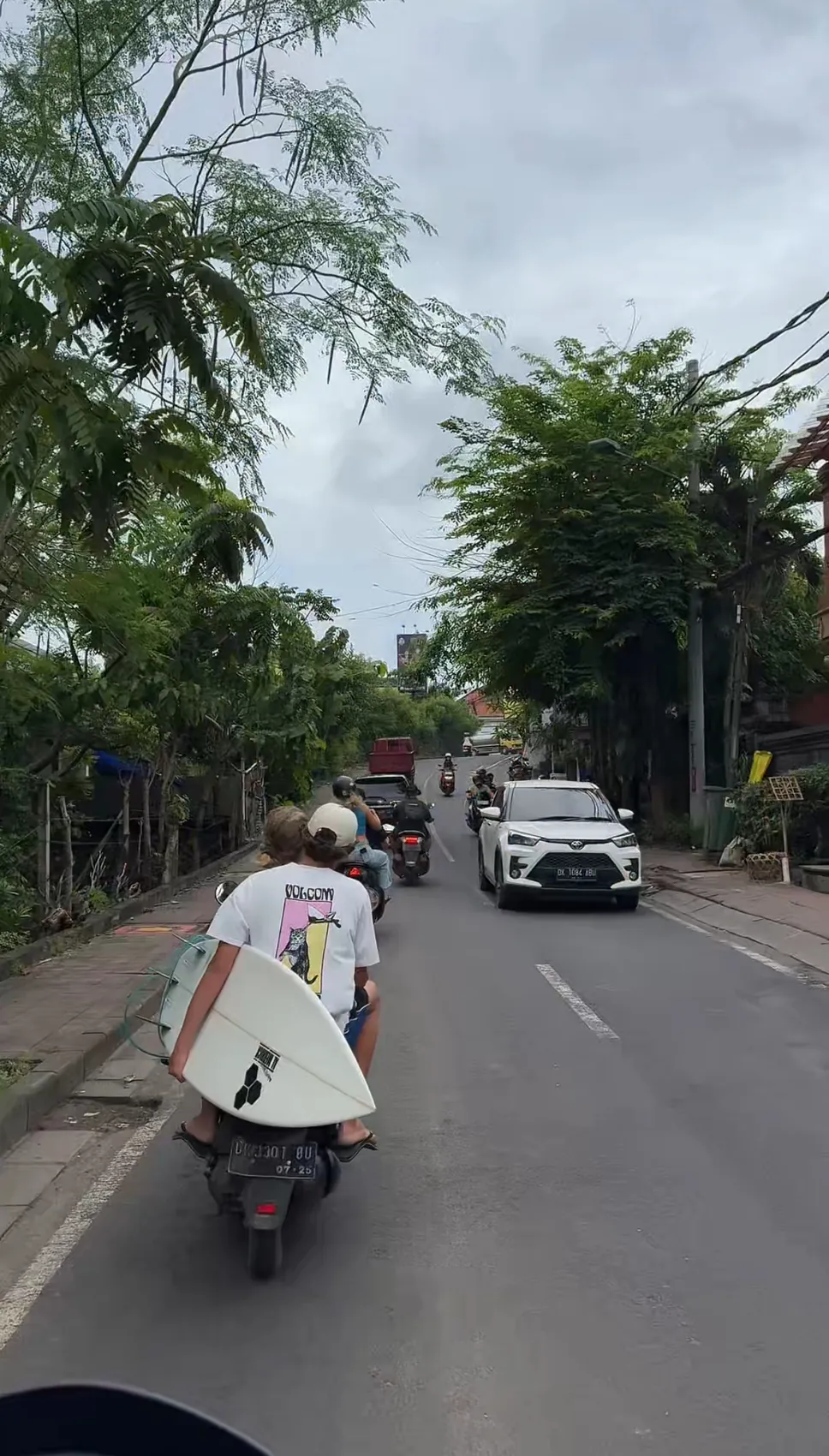A person on a scooter carrying a surfboard down a tree-lined street