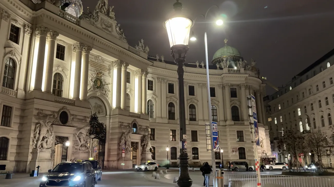 The illuminated Hofburg facade at night with a street lamp and passing taxis.