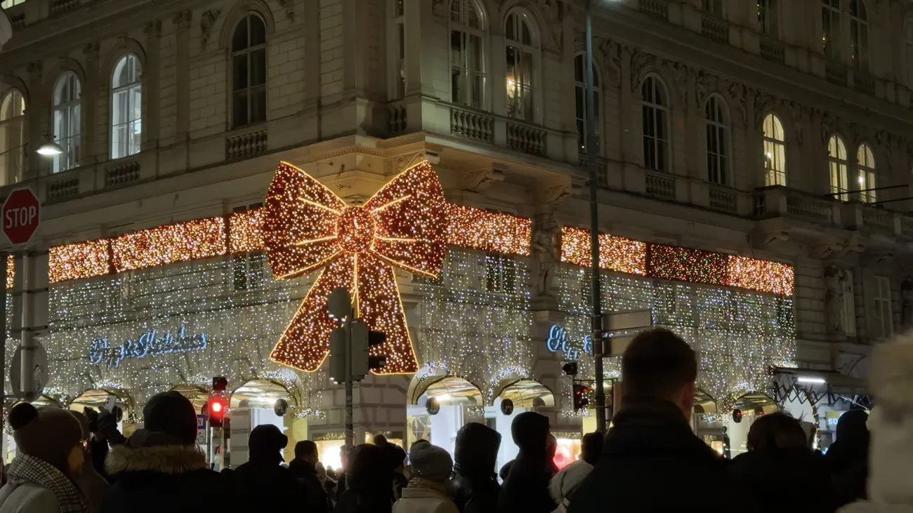 A giant lit-up bow on the corner of a building covered in lights, with a crowd in the foreground.